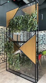 A decorative partition featuring a modern design with geometric wooden panels and metal mesh. It is partially covered with lush green foliage, creating a natural and contemporary look. In the background, there are chairs in various colors, including red, green, and beige, and a tiled wall with a geometric pattern.