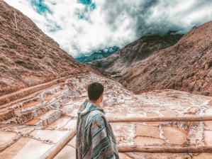 A person wrapped in a colorful blanket stands overlooking a series of terraced salt pans set in a mountainous landscape under a cloudy sky. The terraces are filled with water, displaying various shades of brown and white, nestled between steep, rugged mountains.