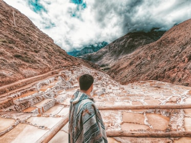 A person wrapped in a colorful blanket stands overlooking a series of terraced salt pans set in a mountainous landscape under a cloudy sky. The terraces are filled with water, displaying various shades of brown and white, nestled between steep, rugged mountains.