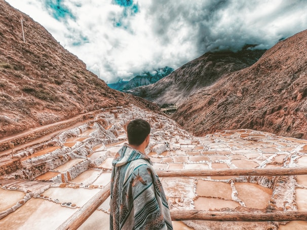 A person wrapped in a colorful blanket stands overlooking a series of terraced salt pans set in a mountainous landscape under a cloudy sky. The terraces are filled with water, displaying various shades of brown and white, nestled between steep, rugged mountains.