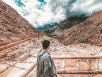 A person wrapped in a colorful blanket stands overlooking a series of terraced salt pans set in a mountainous landscape under a cloudy sky. The terraces are filled with water, displaying various shades of brown and white, nestled between steep, rugged mountains.