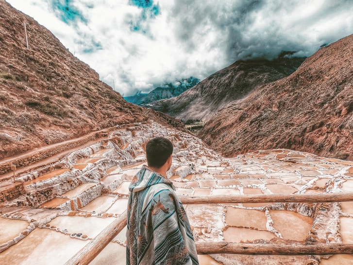 A person wrapped in a colorful blanket stands overlooking a series of terraced salt pans set in a mountainous landscape under a cloudy sky. The terraces are filled with water, displaying various shades of brown and white, nestled between steep, rugged mountains.