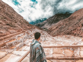 A person wrapped in a colorful blanket stands overlooking a series of terraced salt pans set in a mountainous landscape under a cloudy sky. The terraces are filled with water, displaying various shades of brown and white, nestled between steep, rugged mountains.