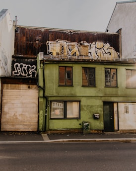 An urban scene featuring a two-story building with a partially worn green facade. The structure has a wooden panel covering part of the second floor. Large graffiti art is prominently displayed on the upper section of the building. Windows on both floors are visible, with some partially boarded or covered. The surrounding area appears gritty, emphasizing an urban, slightly neglected setting.