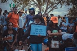 A group of people are gathered outdoors, with some holding signs and others bowing down. A woman in the center holds a sign that reads, 'We are the joy of nations.' The crowd includes individuals of different ages, some standing and others kneeling, and there are trees in the background.