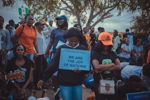 A group of people are gathered outdoors, with some holding signs and others bowing down. A woman in the center holds a sign that reads, 'We are the joy of nations.' The crowd includes individuals of different ages, some standing and others kneeling, and there are trees in the background.