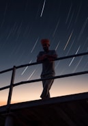 A person wearing a head covering stands on a metal platform, leaning against the railing. The sky is marked with long streaks of light from the stars, indicating a long exposure shot. The horizon is softly lit with tones of orange and blue, suggesting dusk or dawn.
