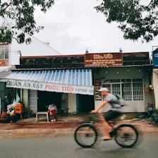 A street scene featuring a small shop with a striped awning and signage. A person sits under the awning, while another person rides a bicycle passing by. The shop appears to be a local eatery with signage in Vietnamese, and the surroundings include trees and other buildings.