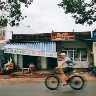 A street scene featuring a small shop with a striped awning and signage. A person sits under the awning, while another person rides a bicycle passing by. The shop appears to be a local eatery with signage in Vietnamese, and the surroundings include trees and other buildings.