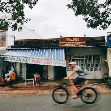 A street scene featuring a small shop with a striped awning and signage. A person sits under the awning, while another person rides a bicycle passing by. The shop appears to be a local eatery with signage in Vietnamese, and the surroundings include trees and other buildings.