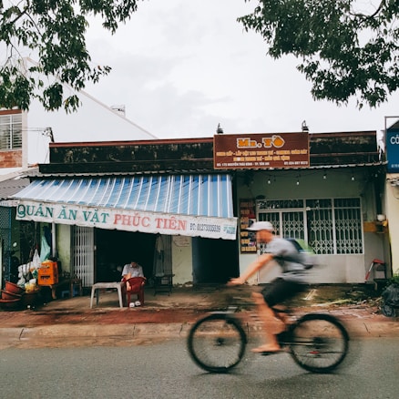 A street scene featuring a small shop with a striped awning and signage. A person sits under the awning, while another person rides a bicycle passing by. The shop appears to be a local eatery with signage in Vietnamese, and the surroundings include trees and other buildings.