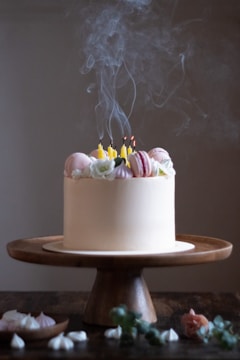 A beautifully decorated white cake on a wooden cake stand features multiple extinguished yellow candles with smoke trailing upwards. The cake is adorned with delicate macarons and floral icing. The setting includes a few small meringues and a pink rose on the table, contributing to an elegant and celebratory atmosphere.
