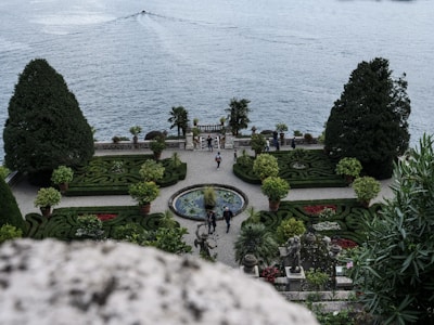 An elegant garden overlooking a large body of water with meticulously trimmed hedges and ornamental plants. The garden features symmetrical designs, a fountain at the center, and statues. There are a few people walking or standing, enjoying the scenery. The location seems serene, with a boat moving in the distance on the water.