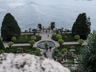 An elegant garden overlooking a large body of water with meticulously trimmed hedges and ornamental plants. The garden features symmetrical designs, a fountain at the center, and statues. There are a few people walking or standing, enjoying the scenery. The location seems serene, with a boat moving in the distance on the water.
