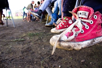 A close-up of worn, red canvas sneakers with white soles and laces on the feet of a child, suspended above the ground. In the background, several people are sitting on chairs arranged in a row, with their legs and feet clearly visible. The ground is dirt and there is a slightly blurred green and beige structure in the distance.