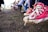 A close-up of worn, red canvas sneakers with white soles and laces on the feet of a child, suspended above the ground. In the background, several people are sitting on chairs arranged in a row, with their legs and feet clearly visible. The ground is dirt and there is a slightly blurred green and beige structure in the distance.