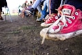 A close-up of worn, red canvas sneakers with white soles and laces on the feet of a child, suspended above the ground. In the background, several people are sitting on chairs arranged in a row, with their legs and feet clearly visible. The ground is dirt and there is a slightly blurred green and beige structure in the distance.