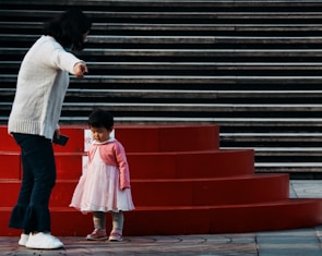 A woman gestures toward a small child in a pink dress standing by red circular steps. The background features a series of dark gray stairs, creating a contrasting backdrop. The woman holds a phone in one hand and appears to be guiding or instructing the child.