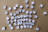 A collection of white six-sided dice scattered randomly on a brown cardboard surface. The dice have black pips, and some display red text on certain sides. The arrangement is haphazard, with various numbers facing upwards.