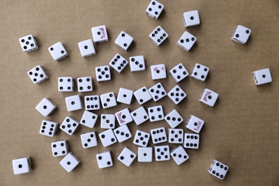 A collection of white six-sided dice scattered randomly on a brown cardboard surface. The dice have black pips, and some display red text on certain sides. The arrangement is haphazard, with various numbers facing upwards.