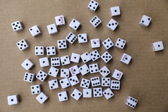 A collection of white six-sided dice scattered randomly on a brown cardboard surface. The dice have black pips, and some display red text on certain sides. The arrangement is haphazard, with various numbers facing upwards.