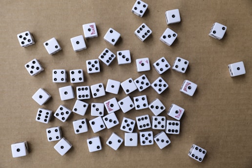 A collection of white six-sided dice scattered randomly on a brown cardboard surface. The dice have black pips, and some display red text on certain sides. The arrangement is haphazard, with various numbers facing upwards.