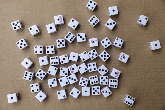 A collection of white six-sided dice scattered randomly on a brown cardboard surface. The dice have black pips, and some display red text on certain sides. The arrangement is haphazard, with various numbers facing upwards.