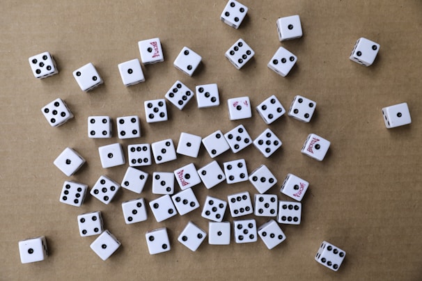 A collection of white six-sided dice scattered randomly on a brown cardboard surface. The dice have black pips, and some display red text on certain sides. The arrangement is haphazard, with various numbers facing upwards.