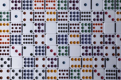 A collection of dominoes is arranged in a grid pattern, each displaying different combinations of colored dots. The dots come in various colors, including orange, purple, yellow, red, green, and blue. The domino pieces are set against a clean, white background.