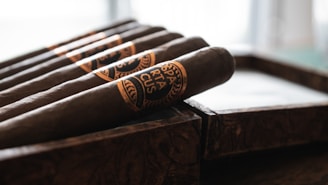 A close-up of several cigars neatly placed in a wooden box. The cigars have a dark wrapper and are adorned with decorative bands featuring intricate designs and text. The background is softly blurred, emphasizing the focus on the cigars.