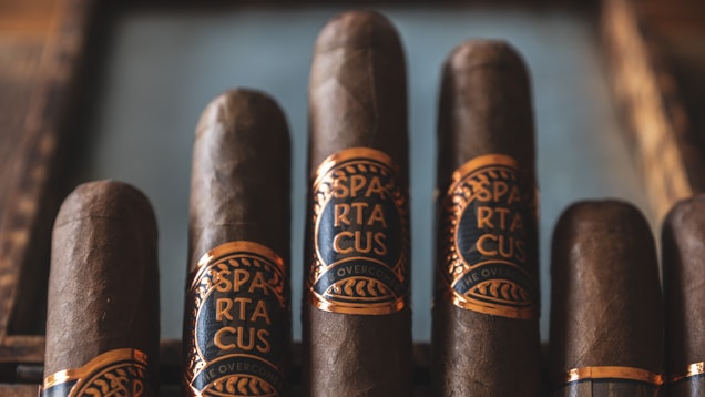 Five cigars are placed in a row, each wrapped with a blue and gold label that reads 'Spartacus'. They have a rich, dark brown coloration, indicating quality tobacco. The setup appears organized and luxurious, suggesting a premium product presentation.