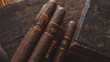 Four cigars with dark brown hues wrapped in ornate bands sit on a rustic wooden surface. The lighting is warm, casting a rich, inviting glow over the scene, complemented by an elegant, engraved wooden box nearby.