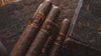 Four cigars with dark brown hues wrapped in ornate bands sit on a rustic wooden surface. The lighting is warm, casting a rich, inviting glow over the scene, complemented by an elegant, engraved wooden box nearby.