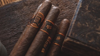 Four cigars with dark brown hues wrapped in ornate bands sit on a rustic wooden surface. The lighting is warm, casting a rich, inviting glow over the scene, complemented by an elegant, engraved wooden box nearby.