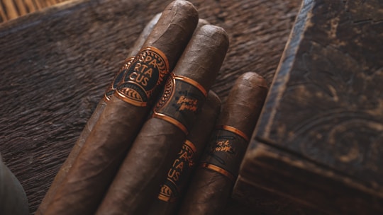 Four cigars with dark brown hues wrapped in ornate bands sit on a rustic wooden surface. The lighting is warm, casting a rich, inviting glow over the scene, complemented by an elegant, engraved wooden box nearby.