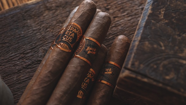 Four cigars with dark brown hues wrapped in ornate bands sit on a rustic wooden surface. The lighting is warm, casting a rich, inviting glow over the scene, complemented by an elegant, engraved wooden box nearby.