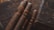 Four cigars with dark brown hues wrapped in ornate bands sit on a rustic wooden surface. The lighting is warm, casting a rich, inviting glow over the scene, complemented by an elegant, engraved wooden box nearby.