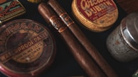 Two cigars placed alongside vintage tins, including one labeled 'Waterproof Paste' and another circular tin labeled 'Dubbin'. The scene has a nostalgic and rustic appearance with a focus on old-fashioned grooming products.