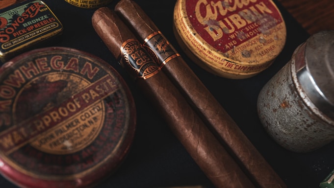 Two cigars placed alongside vintage tins, including one labeled 'Waterproof Paste' and another circular tin labeled 'Dubbin'. The scene has a nostalgic and rustic appearance with a focus on old-fashioned grooming products.