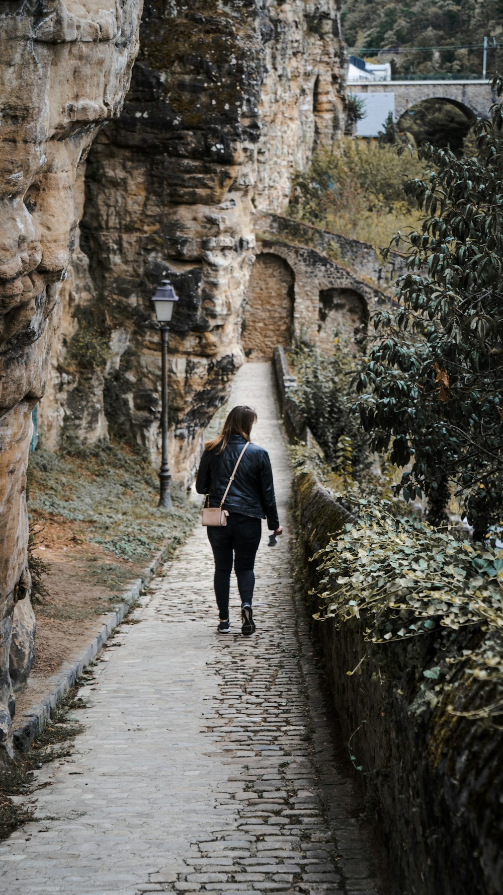 Femme en veste noire marchant sur le sentier entre les murs de béton brun pendant la journée