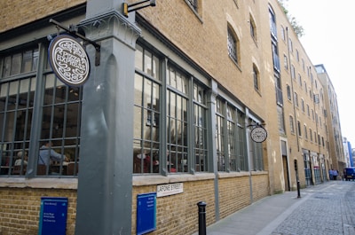 A brick building with large windows stretches along a cobblestone street. There are signs for Pizza Express attached to the building. The scene suggests an urban setting, featuring a quaint street corner with a few people visible in the distance and inside the building.