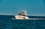 A luxury yacht is cruising through a calm, blue sea, with people on board enjoying the view. The sky is clear, and the ocean stretches into the horizon.