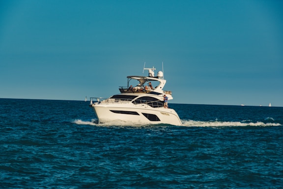 A luxury yacht is cruising through a calm, blue sea, with people on board enjoying the view. The sky is clear, and the ocean stretches into the horizon.