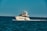 A luxury yacht is cruising through a calm, blue sea, with people on board enjoying the view. The sky is clear, and the ocean stretches into the horizon.