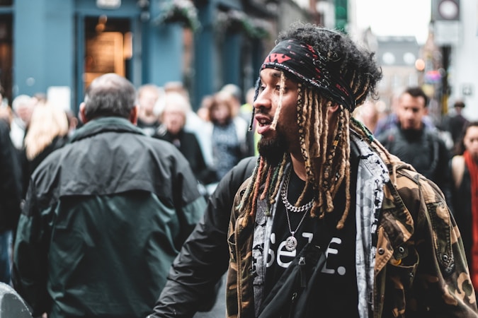 A person with dreadlocks and a bandana walks through a busy street, surrounded by a crowd of people. They wear a camouflage jacket over a graphic t-shirt and accessorize with a chain necklace. The background is slightly blurred, emphasizing the bustling urban environment.