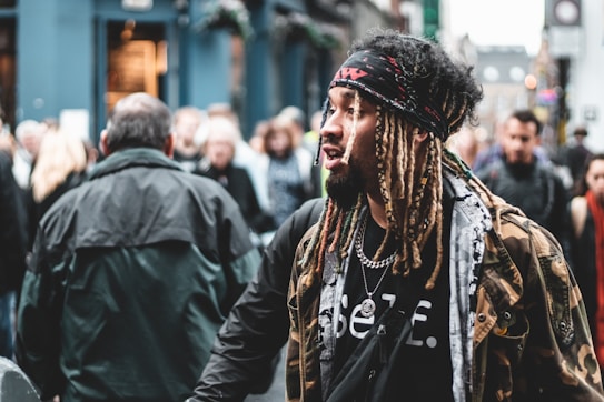A person with dreadlocks and a bandana walks through a busy street, surrounded by a crowd of people. They wear a camouflage jacket over a graphic t-shirt and accessorize with a chain necklace. The background is slightly blurred, emphasizing the bustling urban environment.