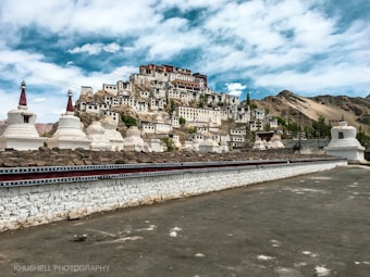 A hillside monastery with a series of white buildings and red roofs is set against a mountainous backdrop. The foreground contains several white stupas and a stone wall. The sky is partly cloudy, enhancing the serene and majestic atmosphere.