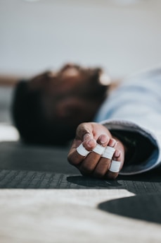 A person is lying on a mat with their fingers wrapped in white athletic tape, indicating an involvement in physical activity or sport. The focus is on their hand, which is extended towards the camera.