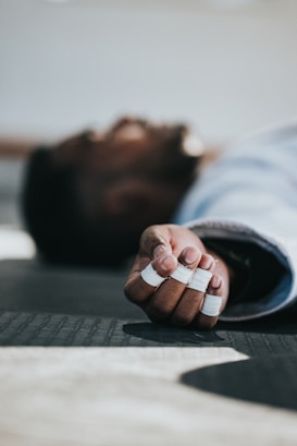 A person is lying on a mat with their fingers wrapped in white athletic tape, indicating an involvement in physical activity or sport. The focus is on their hand, which is extended towards the camera.
