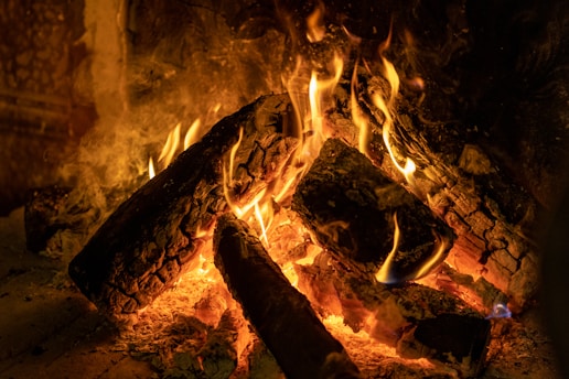 Logs are burning with bright orange and yellow flames in a fireplace. The wood is charred and glowing, surrounded by ashes. The scene emits warmth and coziness.